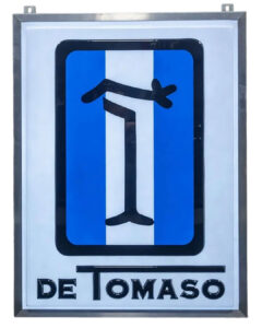 1970s Original De Tomaso Lighted Sign, 48.5”x37”. Metal framed with hanging holes at top. Single-sided white plastic background with blue/white striped De Tomaso large in middle, and the word, De Tomaso at the bottom.